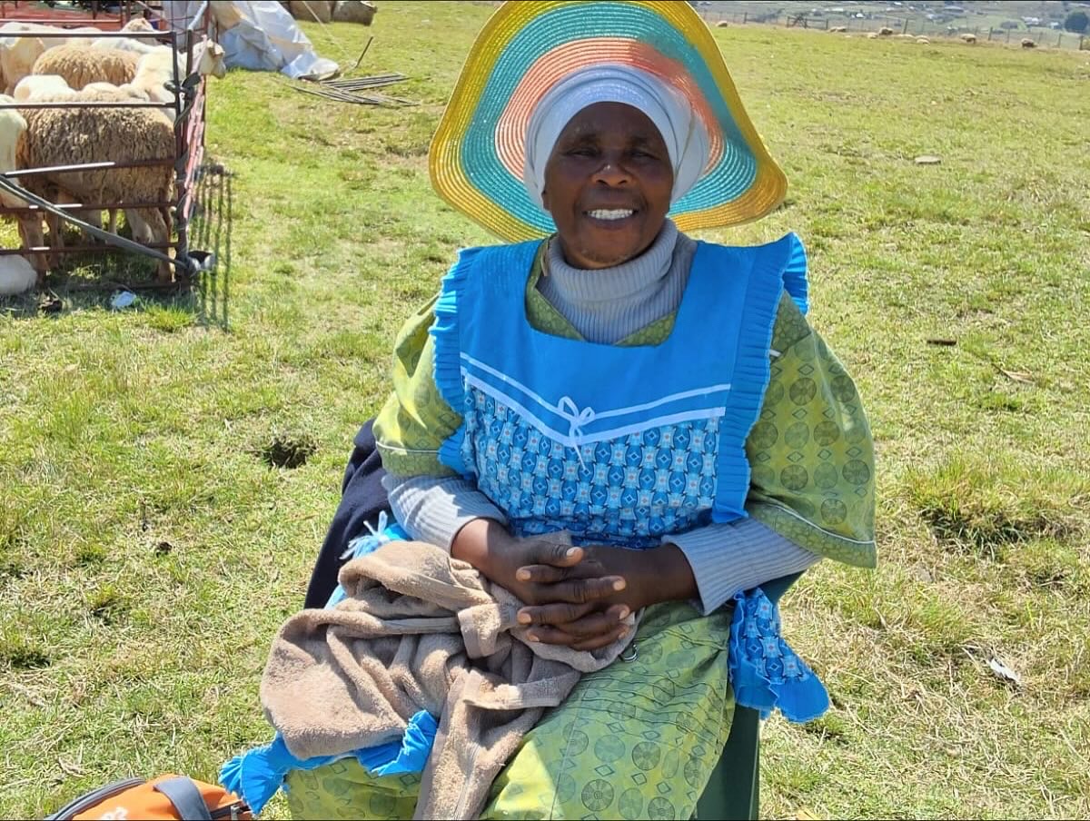 A smiling farmer in a woven hat sits beside a pen of sheep.