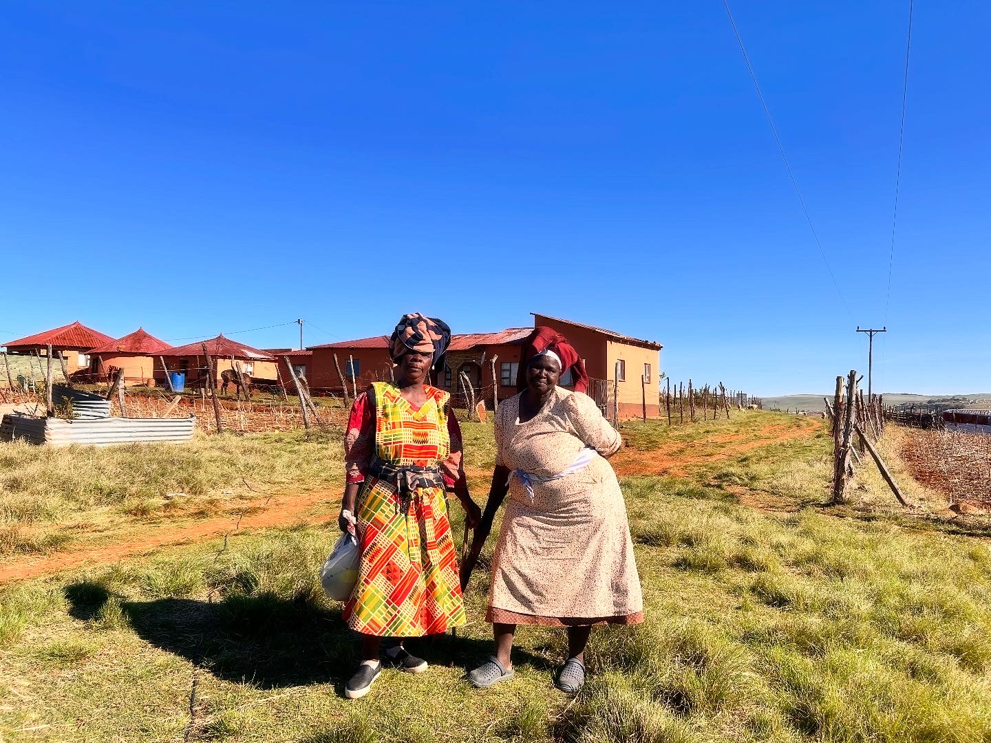 Two women in traditional dress stand together on a farm path near the village.