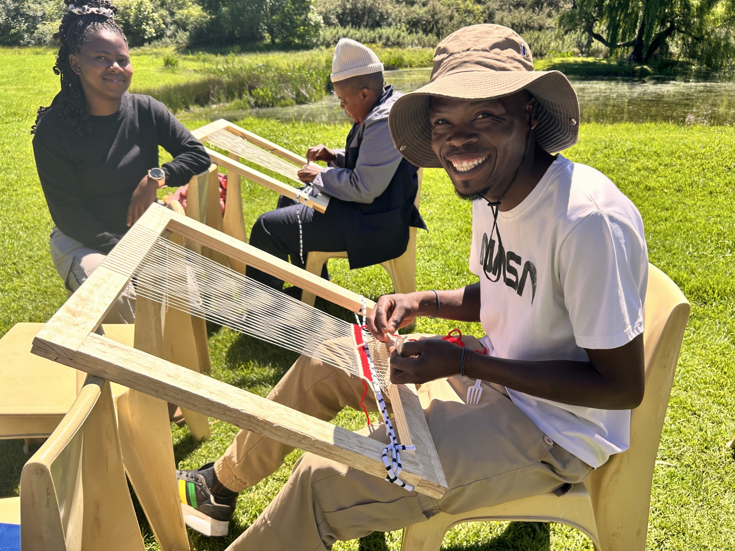 A group of community learners smile while weaving on handmade wooden looms during an outdoor workshop.