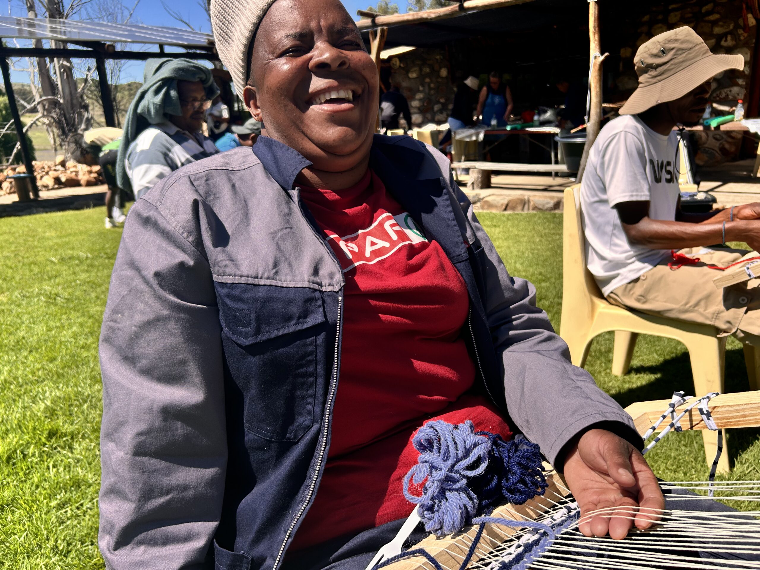 A woman laughs while weaving on a loom, blue wool yarn in hand.